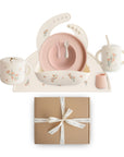 The mushie Deluxe Pink Flowers/Blush Feeding Kit features a floral bib, two-handled cup, bowl with two spoons, sippy cup, plate, and tray—all displayed above a brown gift box tied with a white ribbon.