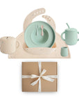The mushie Deluxe Vanilla Confetti/Cambridge Blue Feeding Kit includes a speckled silicone bib, plate, spoon, fork, handled cup, sippy cup, and small container—all toddler feeding essentials in soft pastels presented as a perfect gift.