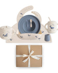 The mushie Deluxe Whales/Tradewinds Feeding Kit includes a blue plate, spoon, fork, two-handled mug, spill-proof cup, and whale placemat neatly arranged above a brown gift box tied with a white ribbon.