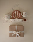 A beige mat showcases the mushie Deluxe Pink Flowers/Blush Feeding Kit—cups, bowls, spoons, and a bib—perfect toddler essentials—arranged above a gift box tied with a white ribbon.