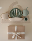 The mushie Deluxe Vanilla Confetti/Cambridge Blue Feeding Kit, featuring a bib, divided plate, bowl, spoons, cup, and sippy cup in soft pastels, is displayed atop a brown gift box with a white ribbon on a cream background.