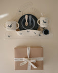 The mushie Deluxe Whales/Tradewinds Feeding Kit, featuring children’s cups, bowls, and spoons with a whale motif, is arranged on a placemat above a brown gift box tied with a white mushie ribbon.