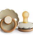 The FRIGG Daisy Natural Rubber Pacifier 2-Pack | Limited Edition features two beige and tan pacifiers with scalloped edges—one standing upright showing the latex nipple, the other flat displaying its ring handle and FRIGG branding.