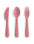 The mushie Dinnerware Cutlery Set features a non-toxic, pink knife, spoon, and fork made from plastic. All pieces are dishwasher-safe and are displayed side by side on a white background.
