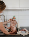 A woman washes a baby in the kitchen sink, gently brushing the baby's head. On the counter, a mushie Suction Spinner Toy spins quietly, offering a soothing sensory experience among the soap bottle and stack of dishes.