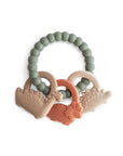 The Dino Teething Ring by mushie is a round, green teething ring made of food-grade silicone with three dinosaur-shaped sensory toys in beige, tan, and orange; the word "mushie" is embossed on the ring.