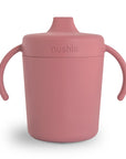The Mushie Trainer Sippy Cup has easy-grip handles, a spill-proof lid, and a pink silicone design with an embossed "mushie" logo for a stylish, child-friendly look.