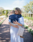 A woman in a white dress hugs a child in a blue sweater on a sunny, flower-lined path, their diaper bag organized with the mushie Water Resistant Wet Bag for easy outings.