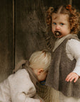 A young girl with curly hair in a dress uses a FRIGG Silicone Pacifier from the FRIGG Silicone Pacifier 6-Pack, standing by a wooden wall, while a blond toddler in a sweater crouches next to her facing downward.
