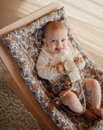 A smiling baby sits in a floral-patterned rocker, holding a bottle fitted with mushie Silicone Bottle Handles. Dressed in cream, the baby is on a cozy rug over a wooden floor, illuminated by soft lighting.