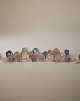 A row of FRIGG Natural Rubber Pacifier 4-Pack pacifiers in soft pastel shades—blue, pink, beige, and brown—are neatly arranged on a neutral backdrop.