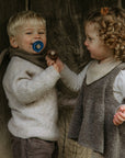 Two children pose before a wooden background; the boy smiles with a blue FRIGG Silicone Pacifier 6-Pack pacifier, while the curly-haired girl on the right reaches out, holding a brown FRIGG pacifier.