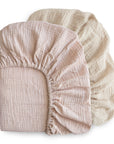 The mushie Extra Soft Muslin Crib Sheet - 2 Pack includes 100% cotton sheets in light pink and cream, featuring elastic edges for a snug fit and a soft, breathable texture.