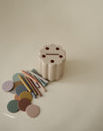 The mushie Coin & Tube Sorting Set, a beige, flower-shaped educational toy with five holes, sits on a light surface surrounded by pastel disks and sticks that help children develop fine motor skills through play.