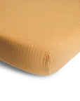 A close-up of the mushie Extra Soft Muslin Crib Sheet in mustard yellow, made from breathable baby bedding, fitted snugly over a mattress corner and displaying its textured fabric against a white background.
