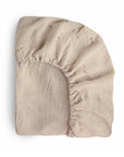 The mushie Extra Soft Muslin Crib Sheet in beige, featuring gentle star and moon patterns and secure elastic edges, is shown partially folded on a white background—ideal for soft, breathable baby bedding.