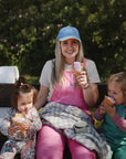 A woman in a blue cap and pink overalls sits on an outdoor bench, smiling and holding ice cream, as two young girls beside her enjoy snacks from mushie Silicone Baby Food Containers. Greenery and flowers frame the scene.