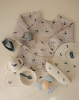 The mushie Long Sleeve Bib is shown with a spoon, bowl, cup, placemat, teether toy, and toy boat in soft blue, grey, and cream with a whale pattern—all neatly arranged on a beige surface.