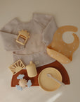 A flat lay of mushie's beige Long Sleeve Bib and other baby essentials, including a wipe-clean bib, plate with spoon, placemat, sippy cup, and teethers—all set on a neutral background.