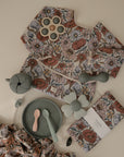 A mushie Long Sleeve Bib, water resistant with a floral pattern and matching cloth, is shown with pastel silicone baby utensils, plate, bear cup, and flower teether on a light background.