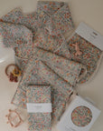A mushie Long Sleeve Bib with a floral print is displayed alongside pastel-colored baby items and visible mushie-branded packaging, all neatly arranged on a light surface.