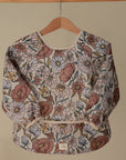The mushie Long Sleeve Bib, featuring a water-resistant fabric and a pink, yellow, and white floral design, hangs on a wooden hanger against a plain beige background.