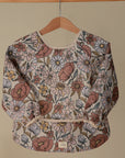The mushie Long Sleeve Bib, featuring a water-resistant fabric and a pink, yellow, and white floral design, hangs on a wooden hanger against a plain beige background.
