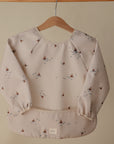 A mushie Long Sleeve Bib in light beige, decorated with small sailboat and wave patterns, is displayed on a wooden hanger against a plain background.