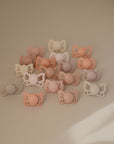 The FRIGG Butterfly Anatomical Silicone Pacifier 2-Pack (6-18 Months) features pastel pacifiers with butterfly-shaped handles, designed for oral development and softly arranged on a beige surface in gentle natural light.