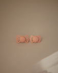 The FRIGG Butterfly Anatomical Silicone Pacifier 2-Pack (0-6 Months) features two peach-colored, bow-shaped pacifiers placed side by side on a beige surface, softly lit to showcase their gentle design for your baby’s comfort.