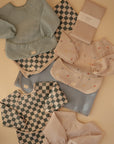 A flat lay on a beige surface featuring Mushie's Splat Mat, pastel and checkered children’s bibs and smocks, and neatly folded fabric pieces.