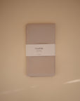 A taupe muslin Mushie Splat Mat, neatly folded and wrapped with a white label featuring the brand name, is displayed against a beige background.