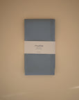 A folded blue Mushie Splat Mat, perfect as a water-resistant high chair mat, is wrapped in a white paper label and set on a plain beige surface.