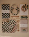 A flat lay of baby essentials from Mushie, including bibs, blankets, teething toys, pacifier clips, utensils, and a water-resistant Splat Mat in muted green, beige, and checkered patterns—all neatly arranged on a light surface.