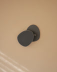 A modern black wall hook is mounted on a smooth beige wall, holding mushie's Cradle Cap Brushes with soft silicone bristles—ideal for baby bath time. Soft diagonal light streaks appear in the lower left corner.