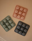 Three Mushie Baby Food Freezer Trays in pastel pink, green, and dark gray, each with nine round molds, are arranged on a beige surface. Ideal for BPA-free baby food storage.