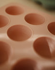 A close-up of the Mushie Baby Food Freezer Tray, a pink BPA-free silicone mold featuring round cups with volume markings in ounces and milliliters, ideal for storing homemade baby food.