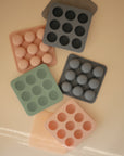 Five Mushie Baby Food Freezer Trays, each pastel-colored and BPA-free, are arranged on a beige surface. Each tray has nine round cavities, some with matching silicone balls, all viewed from above.
