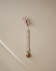 A pink butterfly-shaped pacifier clip hangs on a beige wall, paired with the FRIGG Butterfly Anatomical Silicone Pacifier 2-Pack (6-18 Months). The light strap features a wooden clip, combining charm and comfort from FRIGG for your baby.