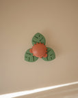The mushie Suction Spinner Toy is a green and orange baby toy made from food-grade silicone, featuring three leaf-shaped sections and a round center, shown on a beige surface with sunlight in the corner.
