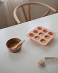 The Mushie Baby Food Freezer Tray, made of pink BPA-free silicone, is shown with yellow batter on a white table next to a matching bowl and spoon, with a wooden chair and baby toy in the background—ideal for storing homemade baby food.