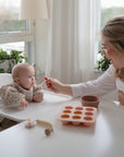 A woman smiles as she feeds a baby in a high chair, who wears the mushie Long Sleeve Bib. Surrounded by plants on the windowsill and a silicone tray with baby food, the cozy room is filled with natural light.