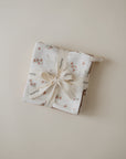 A Muslin Cotton Washcloth by mushie, featuring a folded floral pattern and tied with a cream ribbon printed with the brand name, is displayed on a neutral beige background.