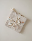 A folded mushie Muslin Cotton Washcloth in beige with small rainbow designs, tied with a white "mushie" ribbon, sits on a plain white surface.