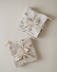Two neatly folded sets of mushie Muslin Cotton Washcloths, one in a floral print and the other in plain beige, tied with branded ribbon and displayed on a cream background.