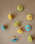 Two FRIGG Rope/Daisy Natural Rubber Pacifiers (Sunflower/Fuchsia, 6-18 Months) for babies are placed on a plain light brown surface. The pacifiers, from FRIGG, feature natural rubber nipples and are baby essentials.