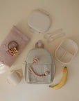 Flat lay of baby items on a light surface: a soft pink blanket with a beaded teether, a beige backpack with the mushie Silicone Pacifier Clip | Luna, a divided lunch container, utensils, lidded cup, and ripe banana.