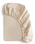 The mushie Stretchy Crib Sheet in beige is shown partly covering a mattress, with its elastic edges gathered at the corner to highlight its stretch and snug fit.