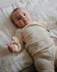 A baby in a cream outfit lies on a white bed, holding the mushie Dog Teether. A pink toy rests nearby as the baby gazes up calmly.