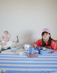A toddler uses a mushie Adjustable Silicone Strap 2-Pack bib while eating at a blue-and-white striped table with an older child in a pink hat slurping noodles nearby.